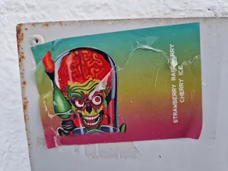 Street sticker A sticker featuring the iconic Mars Attacks alien with a ray gun. The sticker has a colorful, gradient background with text indicating the flavors "Strawberry Raspberry Cherry Ice". The sticker shows some wear and tear from being affixed to a surface.