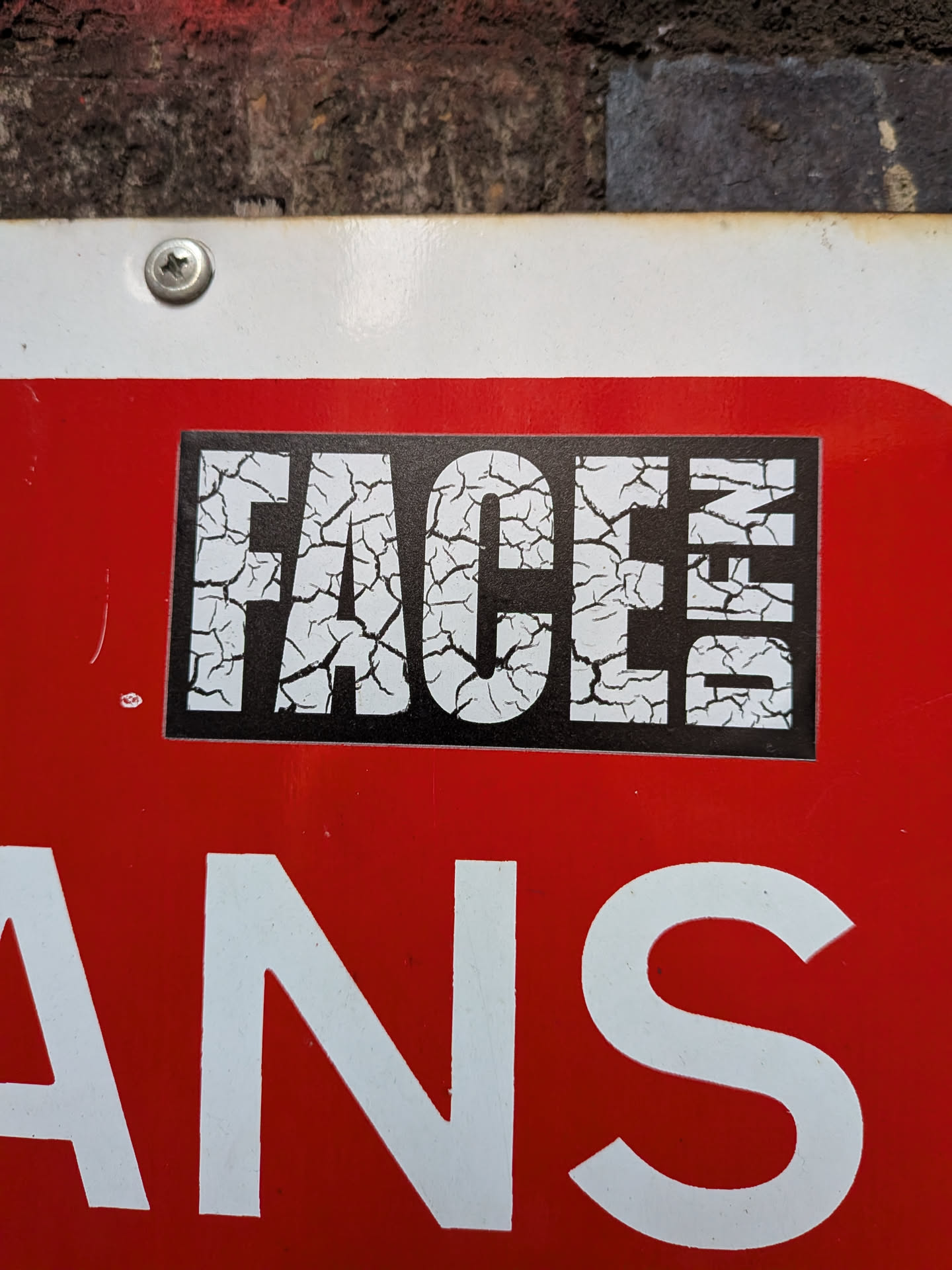 A rectangular black sticker featuring the word 'FACES' in large, uppercase white letters. The white letters have a distressed, cracked texture, appearing as if the surface is breaking apart, revealing black underneath. The sticker is placed centrally on a larger red surface, creating a strong visual contrast.