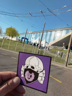 Street sticker The sticker is square-shaped with a thin black border, set against a vibrant purple background. It depicts a stylized white skull, seen from the front, with visible sutures on its cranium. The lower part of the skull, including the jaw, is covered by a black bandana or mask. Centered on this black mask is a purple crest, which is a stylized rendition of the Real Madrid C.F. logo, featuring intertwined letters 'R', 'M', 'C', 'F' within a laurel wreath and topped with a small crown. Two white, bone-like shapes extend from behind the skull on either side, adding to the macabre aesthetic. The overall design is graphic and bold.