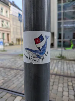 Street sticker The sticker depicts a blue whale, illustrated in a friendly, cartoonish style. Perched on its back is a small figure holding a flag that is predominantly red and blue with some white details. Curving along the bottom edge of the whale, the text "Sandefjord på tur" is written in a black, informal script. The sticker is placed on a textured, dark grey pole, with a blurred background showing urban elements like buildings and cobblestone pavement.