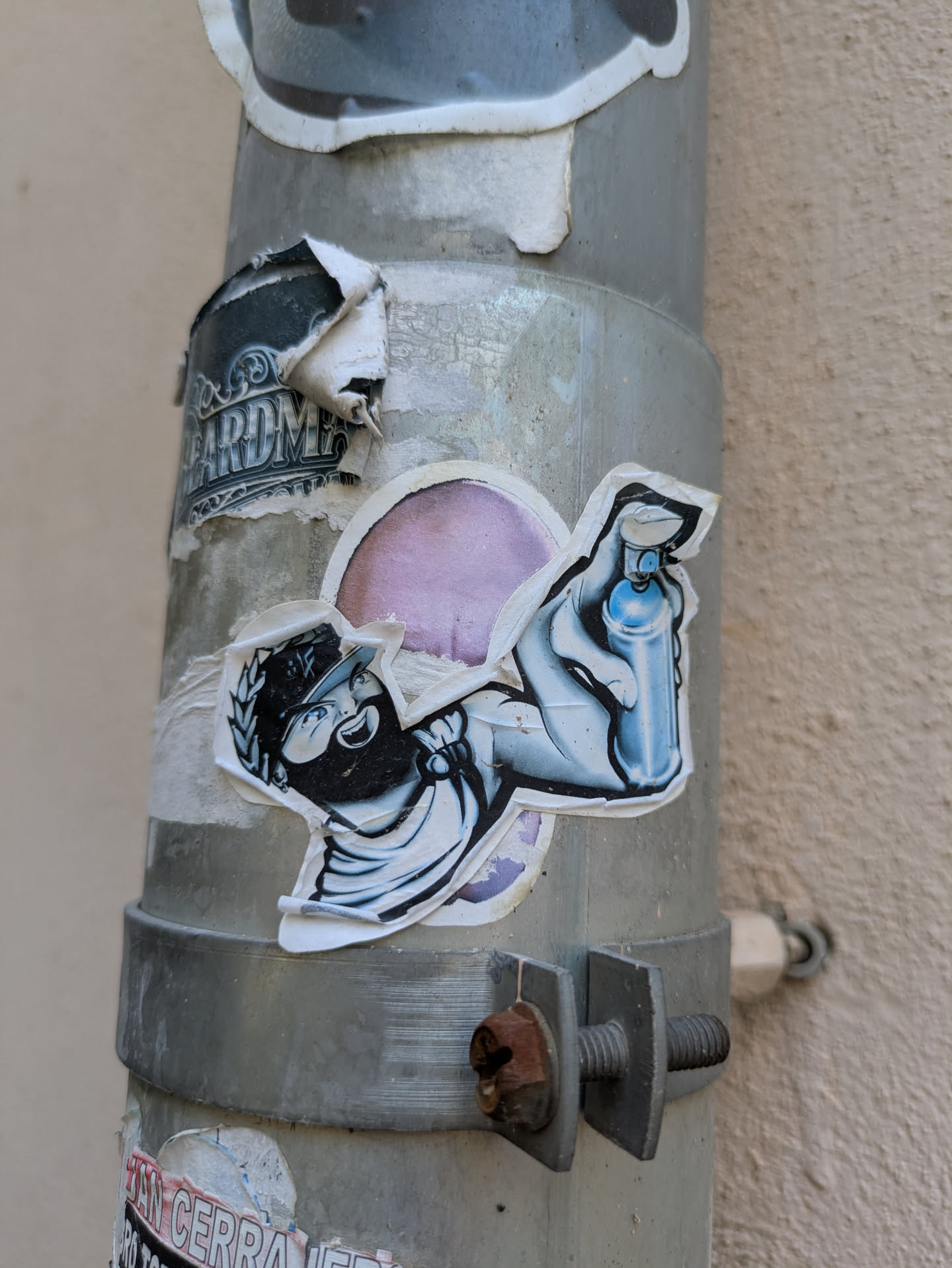 The sticker features a figure with a mask, wearing a hat with a laurel, holding a spray paint can with a backdrop of a round purple spot. The sticker is applied on a metallic pipe alongside other weathered stickers.