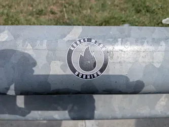 Street sticker A round black sticker on a metal rail. It features a central white-outlined flame icon surrounded by a circular border containing the text 'KORNY WOOD BURNING' in white.