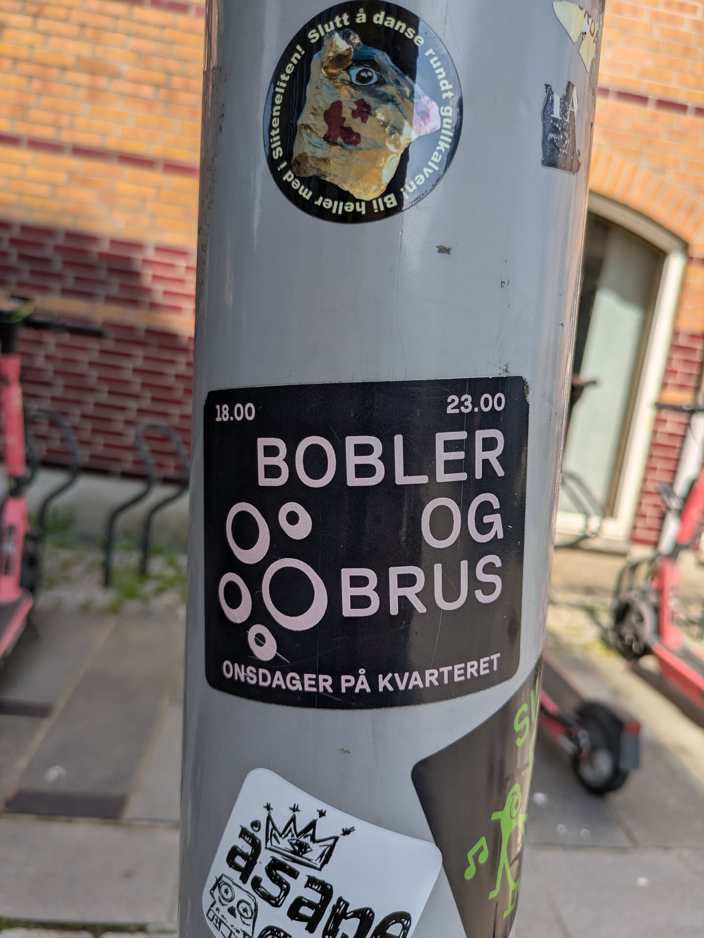 The sticker is rectangular with a black background and features white, sans-serif text. The main text 'BOBLER OG BRUS' is prominently displayed, with stylized white circles of varying sizes between 'BOBLER' and 'OG BRUS' that resemble bubbles. Above the main title, the times '18.00' and '23.00' are placed on the left and right, respectively. Below the main title, the text 'ONSDAGER PÅ KVARTERET' specifies the day and venue of the event.