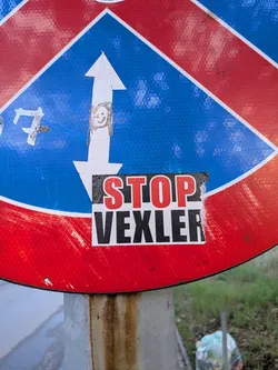 Street sticker Rectangular sticker with two horizontal blocks. Top block has red 'STOP' text on black; bottom block has black 'VEXLER' text on white. Placed on a blue and red road sign.