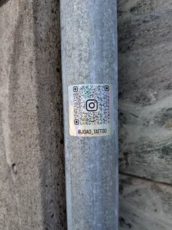 Square holographic sticker on a metal pipe. Features a QR code with an Instagram logo in the center and the handle '@JOAO_TATTOO' printed below in black.