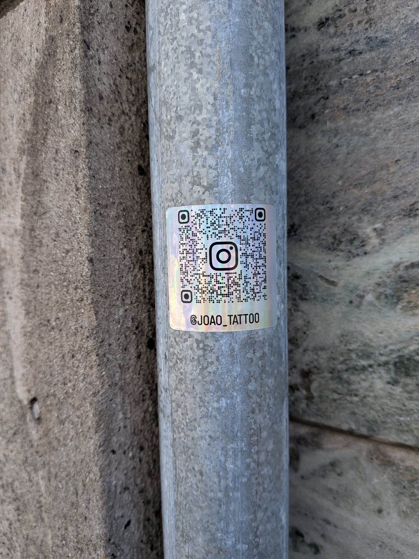 Square holographic sticker on a metal pipe. Features a QR code with an Instagram logo in the center and the handle '@JOAO_TATTOO' printed below in black.