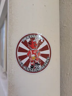 Street sticker The sticker features a red devil mascot, a common symbol for the 1. FC Kaiserslautern football club. The devil is wearing the team's jersey with the number 9, holding a stein of beer in one hand and a soccer ball under its foot. The sticker is circular with red and white stripes in the background and text indicating it's a fan representation. "DIE FANS VOM 1.FC KAISERSLAUTERN".