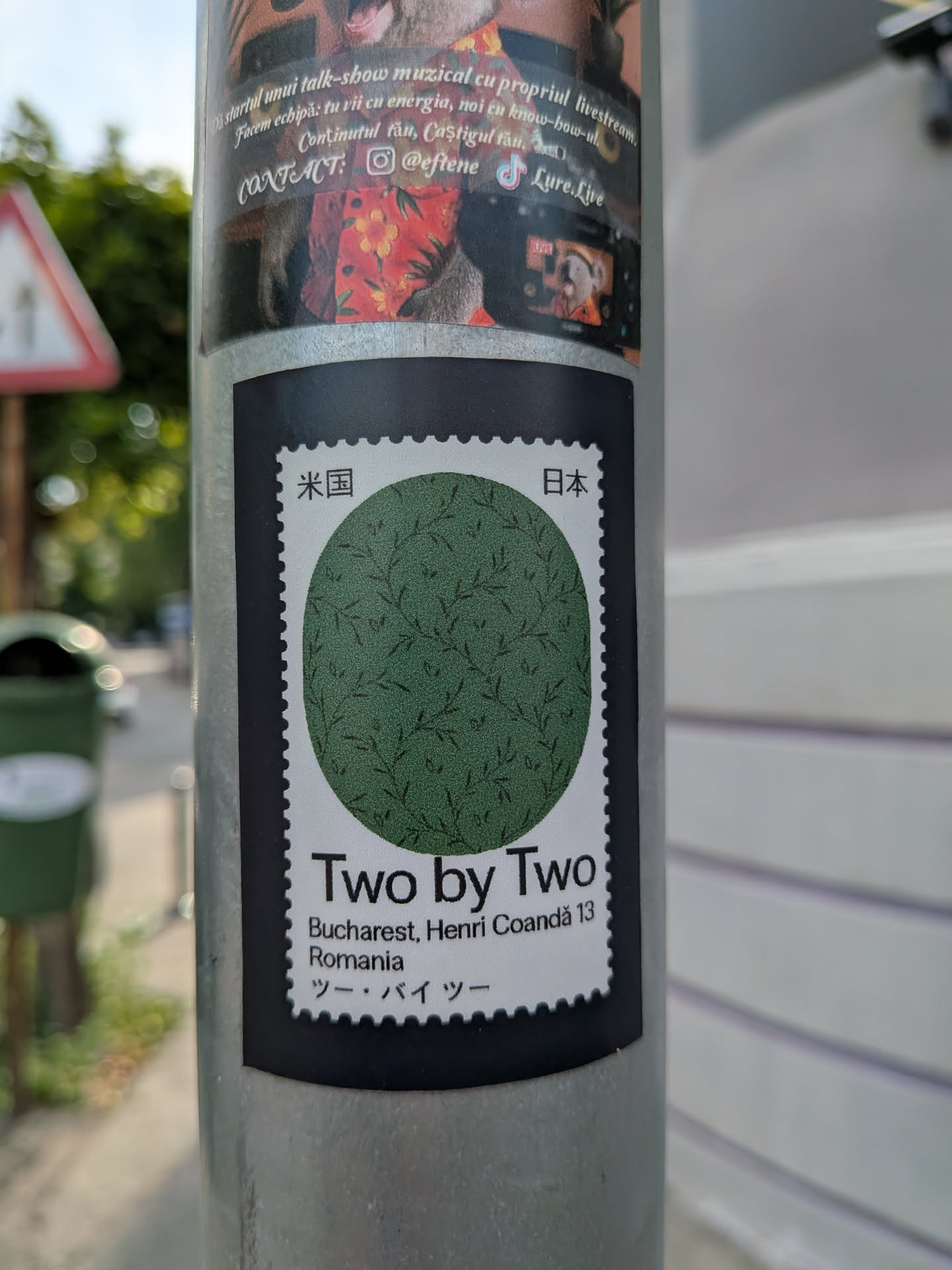 A rectangular sticker styled as a postage stamp with a black outer border. It features a central green circle with a delicate leaf pattern. The top corners display Japanese kanji, while the bottom section contains the text 'Two by Two', a Bucharest address, and its Japanese katakana transliteration.