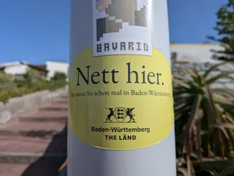 Street sticker An oval yellow sticker featuring the German text 'Nett hier. Aber waren Sie schon mal in Baden-Württemberg?' and the state's coat of arms with 'THE LÄND' branding.