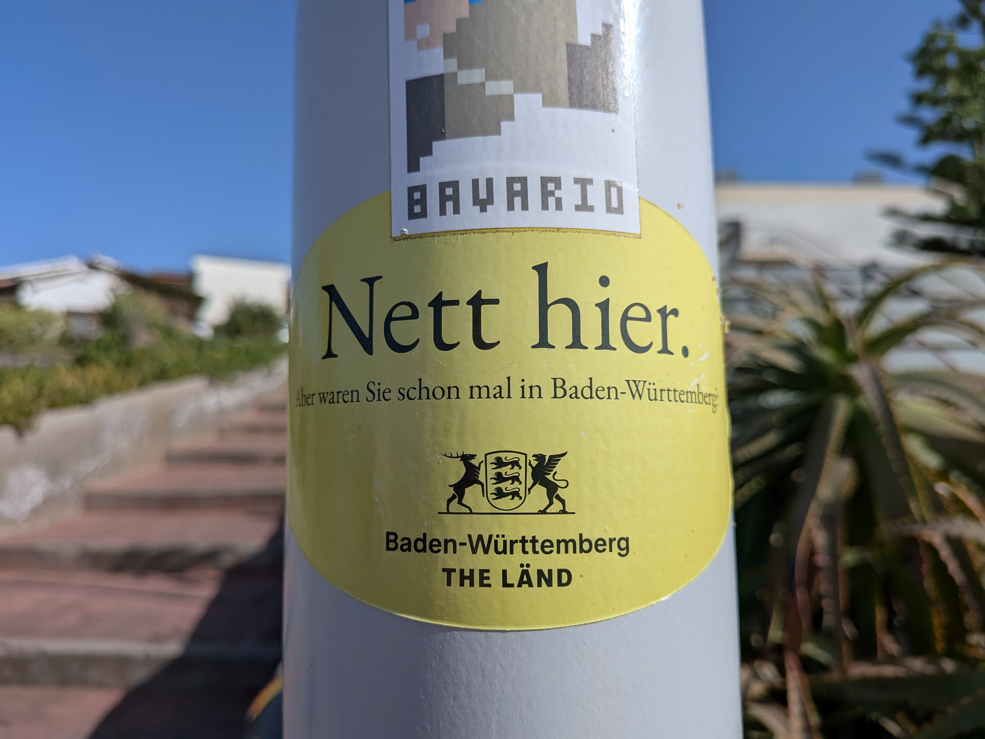 An oval yellow sticker featuring the German text 'Nett hier. Aber waren Sie schon mal in Baden-Württemberg?' and the state's coat of arms with 'THE LÄND' branding.