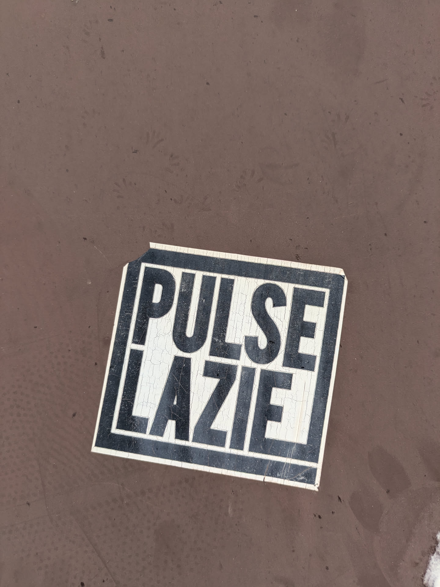 The sticker, featuring a black outline, is slightly torn. The text 'PULSE LAZIE' is prominently displayed. The sticker has a cracked texture, suggesting it's been exposed to the elements for a while. It's placed against a brown surface.