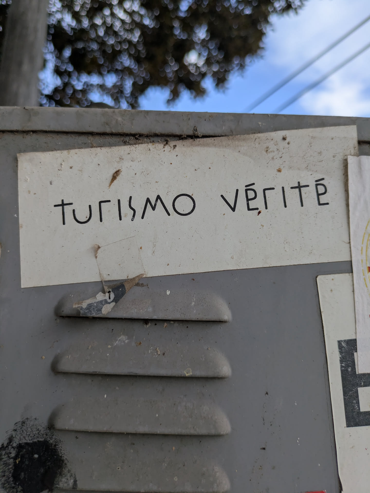 The sticker features the words "Turismo Vérité" in black, printed on a white background, affixed to a gray metal surface. The style of the text is modern and somewhat minimalist. The sticker shows some wear and tear, with small imperfections adding to its character.
