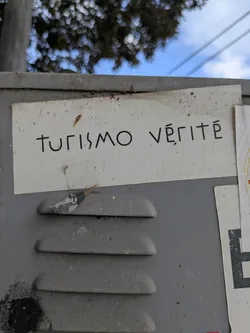 Street sticker The sticker features the words "Turismo Vérité" in black, printed on a white background, affixed to a gray metal surface. The style of the text is modern and somewhat minimalist. The sticker shows some wear and tear, with small imperfections adding to its character.