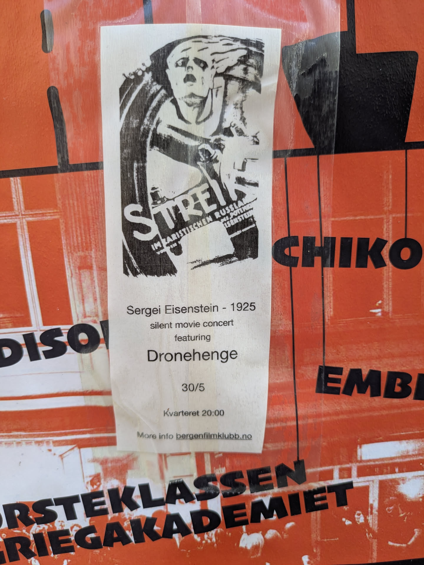 The sticker is a white, vertical rectangle with black print, adhered to a larger orange background. The top half features a striking black and white illustration, likely a poster for the film 'Strike' (Streik), depicting a dramatic, distressed figure. Below the illustration, text provides details about a 'silent movie concert' featuring Sergei Eisenstein's 1925 film, with music by 'Dronehenge'. The date '30/5' and time 'Kvarteret 20:00' are listed, along with a website for more information.