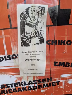 Street sticker The sticker is a white, vertical rectangle with black print, adhered to a larger orange background. The top half features a striking black and white illustration, likely a poster for the film 'Strike' (Streik), depicting a dramatic, distressed figure. Below the illustration, text provides details about a 'silent movie concert' featuring Sergei Eisenstein's 1925 film, with music by 'Dronehenge'. The date '30/5' and time 'Kvarteret 20:00' are listed, along with a website for more information.