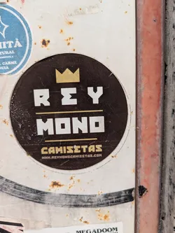 Street sticker The sticker is round, with a dark brown background and a white border. At the top is a golden crown. Below the crown, in white block letters, is the text "REY MONO". Underneath that is the word "CAMISETAS" in gold letters. At the bottom of the sticker is the URL "WWW.REYMONOCAMISTAS.COM" in white letters.
