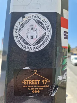 Street sticker A dark brown sticker with the text STREET 17, the address Via G. Vacca, 17 - Monopoli (Ba), and a phone number. It also features a hanger icon and social media icons (Facebook, Instagram, possibly others).  There's a partially visible sticker above it, which appears to be a round emblem with a castle image and text suggesting a barricade or similar theme in Italian. 