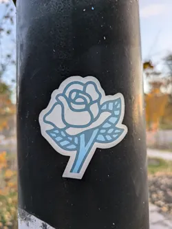 Street sticker A simple sticker of a light blue rose on a dark pole. The rose is depicted in a minimalist style with a few leaves. The sticker appears to be made of vinyl.