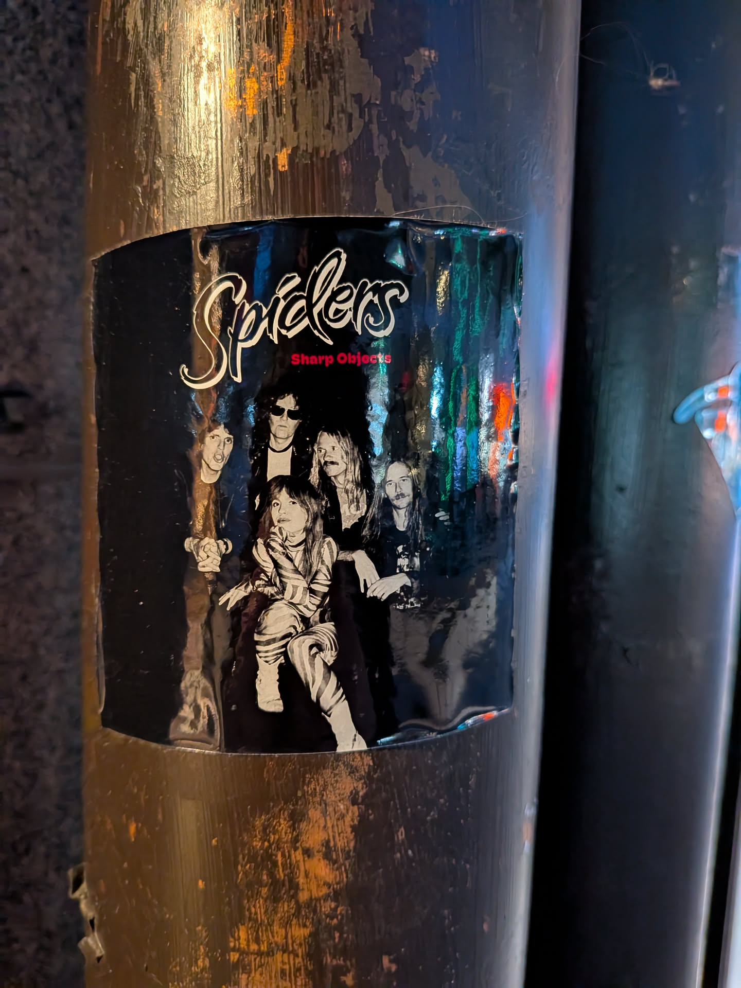 The sticker is black with a white, stylized font spelling out 'Spiders' at the top. Below this, in red text, is 'Sharp Objects'. The main visual is a black and white photograph of a band with five members. One member is seated in the foreground wearing a striped outfit, while the others are standing behind. The background of the photograph appears to be a blurry, colorful urban scene with reflections of lights.