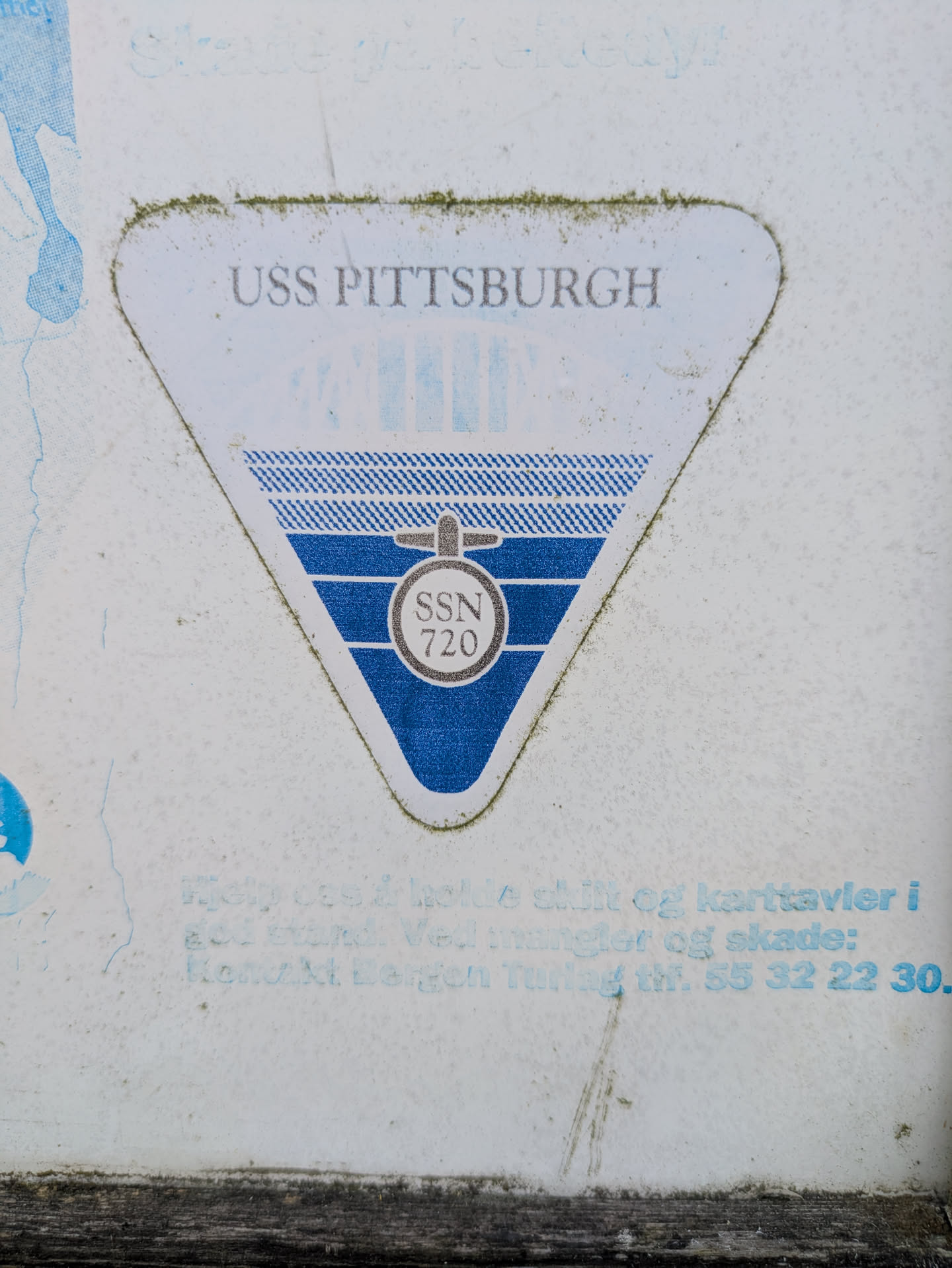 The image displays a white triangular sticker with blue and black accents, affixed to a textured, light-colored surface. At the top of the sticker, the words "USS PITTSBURGH" are prominently displayed in a greyish-blue font. Below this, a stylized emblem in blue depicts what appears to be a ship's sail or a propeller, with horizontal lines. Centered within this design is a circle containing the text "SSN 720" in black. The sticker shows signs of age and exposure, with some dirt and green residue along its edges.
