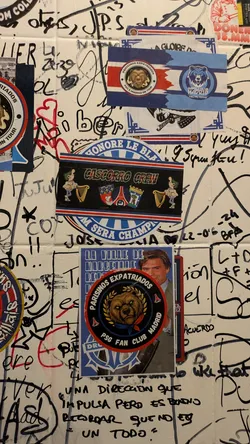 Street sticker A horizontal rectangular sticker featuring a central black band with the text 'CASCORRO CREW' in a stylized, distressed white font with a green outline. To the left of the text is a cartoon character with green hair, wearing a light blue shirt and dark pants, holding a book and playing a golden harp. To the right are two shield-shaped crests; one clearly depicts a fleur-de-lis and possibly the Eiffel Tower, while the other is less distinct. The black band is framed by thin white lines, then red and blue stripes. The top blue stripe partially displays 'HONORE LE BLANC', and the bottom blue stripe partially displays 'M SERA CHAMPAGNE'. The outermost edges feature a decorative blue and white pattern.