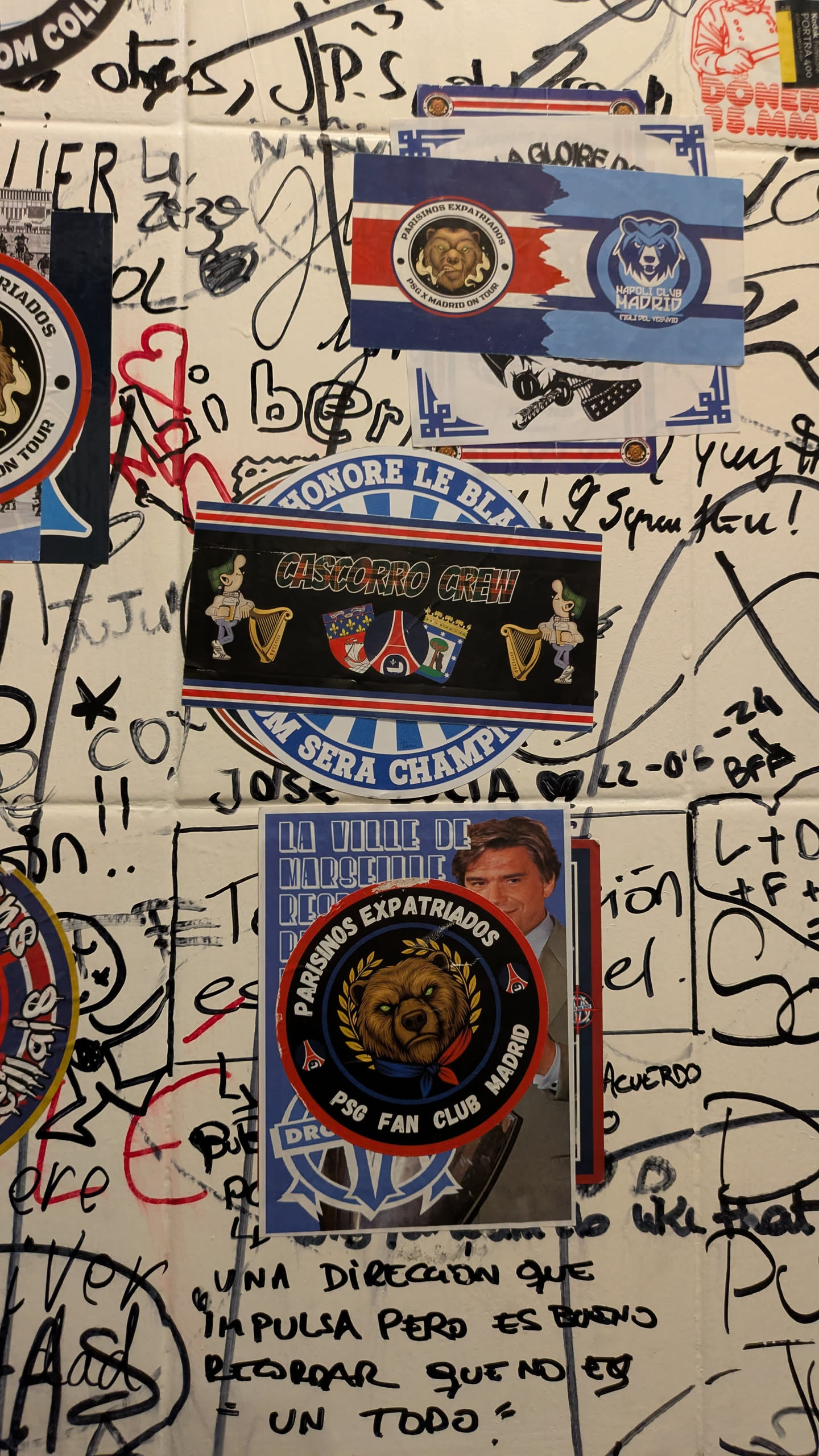 A horizontal rectangular sticker featuring a central black band with the text 'CASCORRO CREW' in a stylized, distressed white font with a green outline. To the left of the text is a cartoon character with green hair, wearing a light blue shirt and dark pants, holding a book and playing a golden harp. To the right are two shield-shaped crests; one clearly depicts a fleur-de-lis and possibly the Eiffel Tower, while the other is less distinct. The black band is framed by thin white lines, then red and blue stripes. The top blue stripe partially displays 'HONORE LE BLANC', and the bottom blue stripe partially displays 'M SERA CHAMPAGNE'. The outermost edges feature a decorative blue and white pattern.