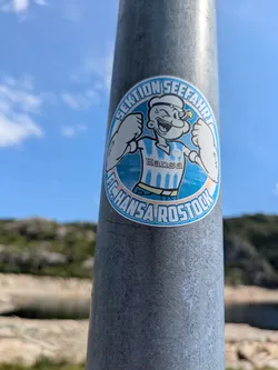 Street sticker A circular sticker affixed to a metal pole, featuring Popeye the Sailor Man. Popeye is depicted with a wide grin, flexing his biceps, and wearing a blue and white striped soccer jersey with "Hansa" written on it. The sticker has a light blue background and is framed by text. The top arc reads "SEKTION SEEFAHRT", and the bottom arc reads "F.C. HANSA ROSTOCK". The background is a bright blue sky with some clouds and blurred distant land.