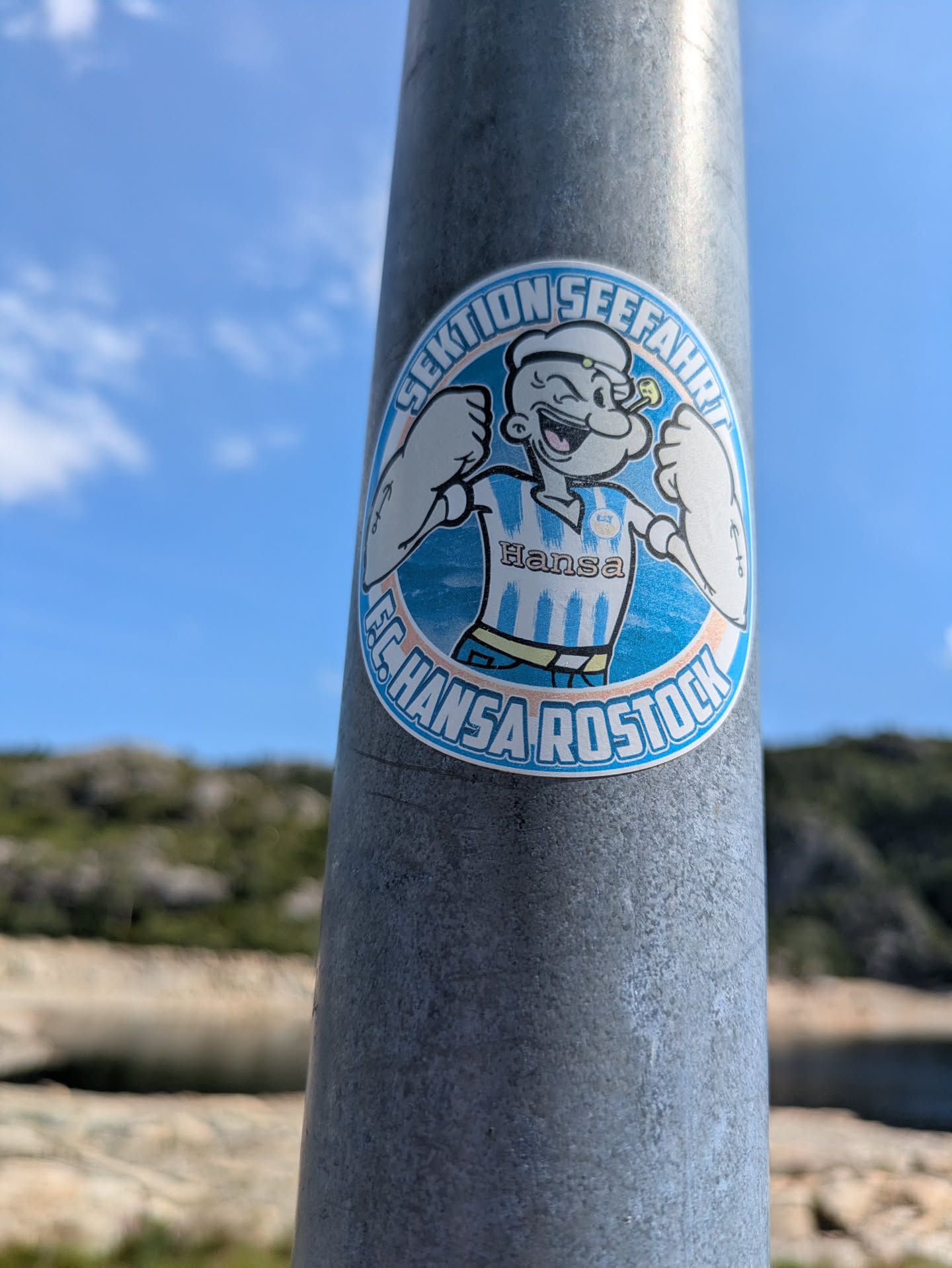 Street sticker A circular sticker affixed to a metal pole, featuring Popeye the Sailor Man. Popeye is depicted with a wide grin, flexing his biceps, and wearing a blue and white striped soccer jersey with "Hansa" written on it. The sticker has a light blue background and is framed by text. The top arc reads "SEKTION SEEFAHRT", and the bottom arc reads "F.C. HANSA ROSTOCK". The background is a bright blue sky with some clouds and blurred distant land.