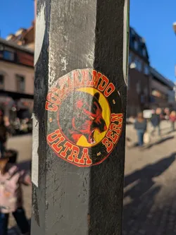 Street sticker This sticker is on a pole. It features a bear logo with a crescent moon behind it, with the words "Commando Ultra Bern" encircling the image. The color scheme is primarily red, yellow, and black.