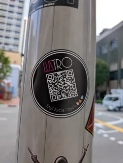 Street sticker A circular black sticker on a metal pole featuring the word 'LUSTRO' in pink and white, a central QR code, and the Polish phrase 'Styl życia i bycia' at the bottom.