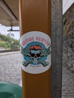 Street sticker Circular sticker depicting a blue skull with spiky orange hair and sunglasses. Behind it are crossed green bones with hands making the 'rock on' sign. Text reads 'HEILIGE GEISTER', 'SÜDKURVE SEIT 2008', and 'FC ST. PAULI'.