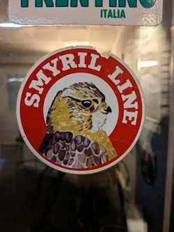 Street sticker The sticker is circular with a red border. Inside the border, white text reads 'SMYRL LINE' in an arc. The center of the sticker features a detailed illustration of a falcon's head and upper chest. The falcon has sharp, dark eyes, a hooked beak, and its plumage is depicted with intricate patterns of yellow, brown, and white. The background within the circle is white, providing a stark contrast to the detailed bird.