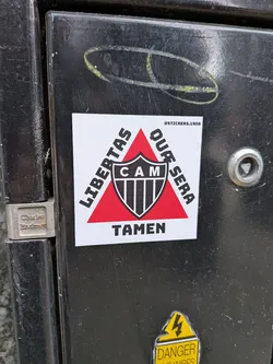 A square white sticker featuring a prominent red triangle pointing upwards. Centered within the triangle is the shield logo of Clube Atlético Mineiro, displaying 'CAM' in white letters on a black background at the top, followed by vertical black and white stripes. Around the red triangle, in bold black capital letters, the Latin motto 'LIBERTAS QUAE SERA TAMEN' is inscribed: 'LIBERTAS' runs diagonally up the left side, 'QUAE SERA' diagonally down the right side, and 'TAMEN' horizontally across the bottom. In the top right corner of the white background, a small black text reads '@STICKERS.1908'. The sticker has a clean, graphic design with a strong contrast between the red, black, and white elements.