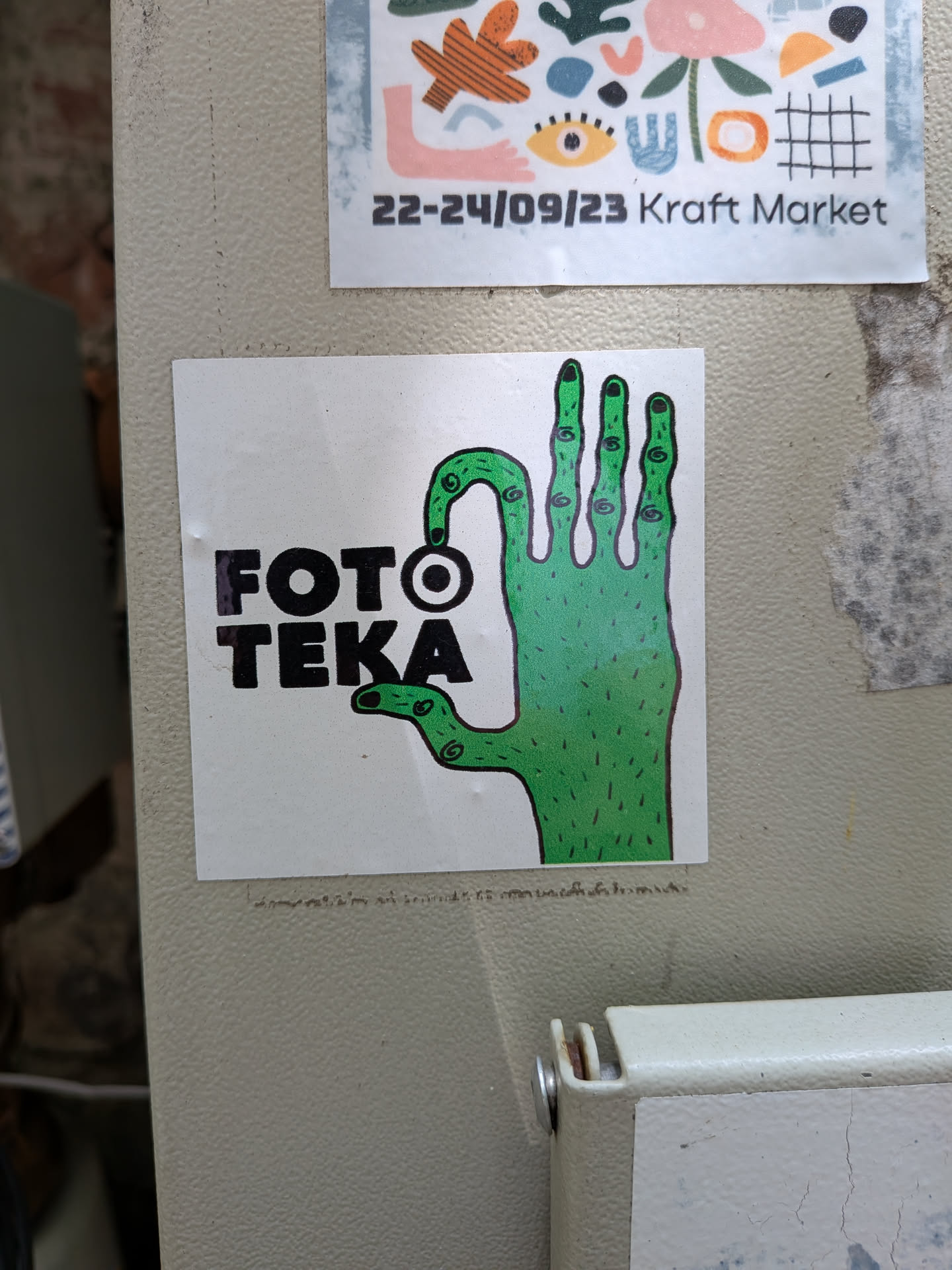 A square white sticker depicting a green, hairy hand with spiral-patterned fingers. The hand is positioned next to the bold black text 'FOTO TEKA', with one finger curving towards the letter 'O'.
