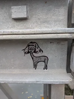 Street sticker The sticker features a black and white illustration of a creature. It has a disproportionately large head with wide, staring eyes and a small mouth. Its hair is depicted as wild and flowing. The body is slender and covered in horizontal stripes, resembling a zebra's pattern. It has four thin legs and appears to be standing. The sticker has a glossy finish and is adhered to a metallic surface.