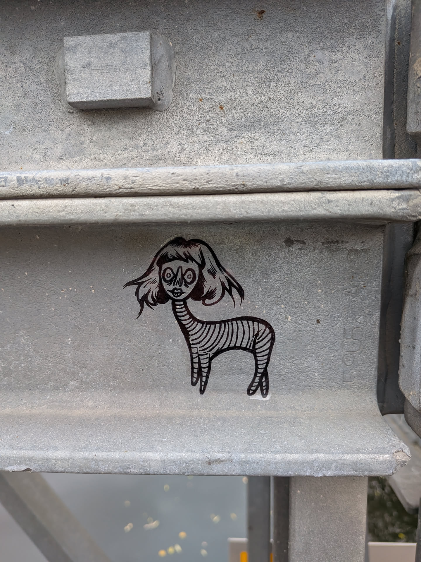 Street sticker The sticker features a black and white illustration of a creature. It has a disproportionately large head with wide, staring eyes and a small mouth. Its hair is depicted as wild and flowing. The body is slender and covered in horizontal stripes, resembling a zebra's pattern. It has four thin legs and appears to be standing. The sticker has a glossy finish and is adhered to a metallic surface.