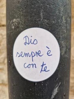 A round white sticker with a handwritten message in blue ink. The message reads: "Dio sempre è con te." which translates from Italian to: "God is always with you."