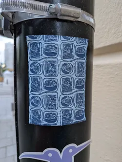 Street sticker A rectangular sticker on a black pole featuring a repeating grid of blue and white map-like illustrations. The text 'FLORAS KULLE MASSIVE' is repeated within the design.