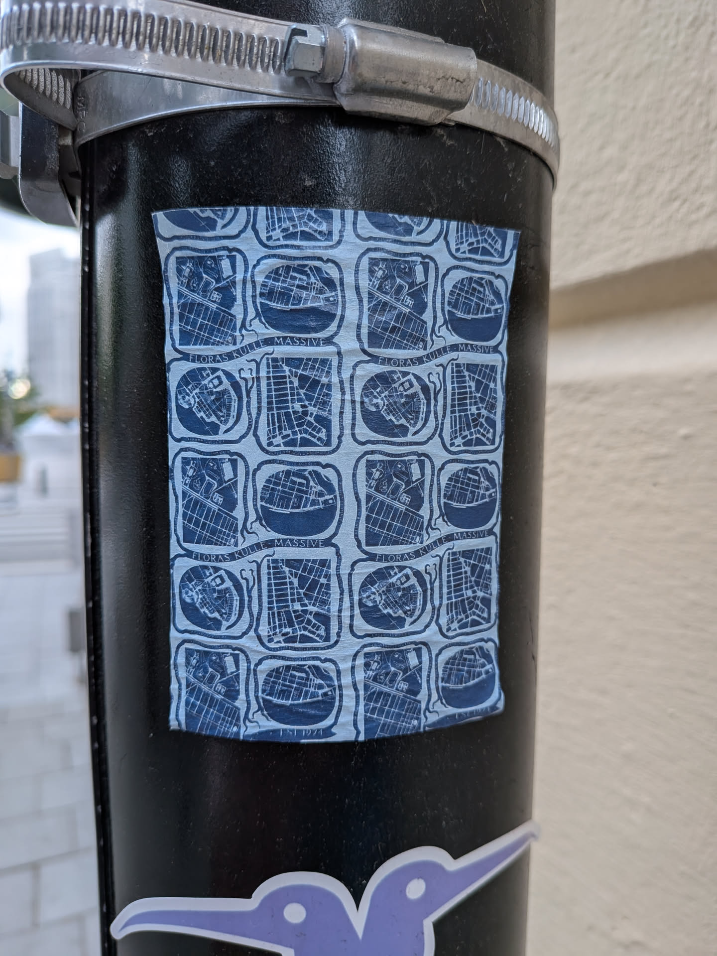 A rectangular sticker on a black pole featuring a repeating grid of blue and white map-like illustrations. The text 'FLORAS KULLE MASSIVE' is repeated within the design.