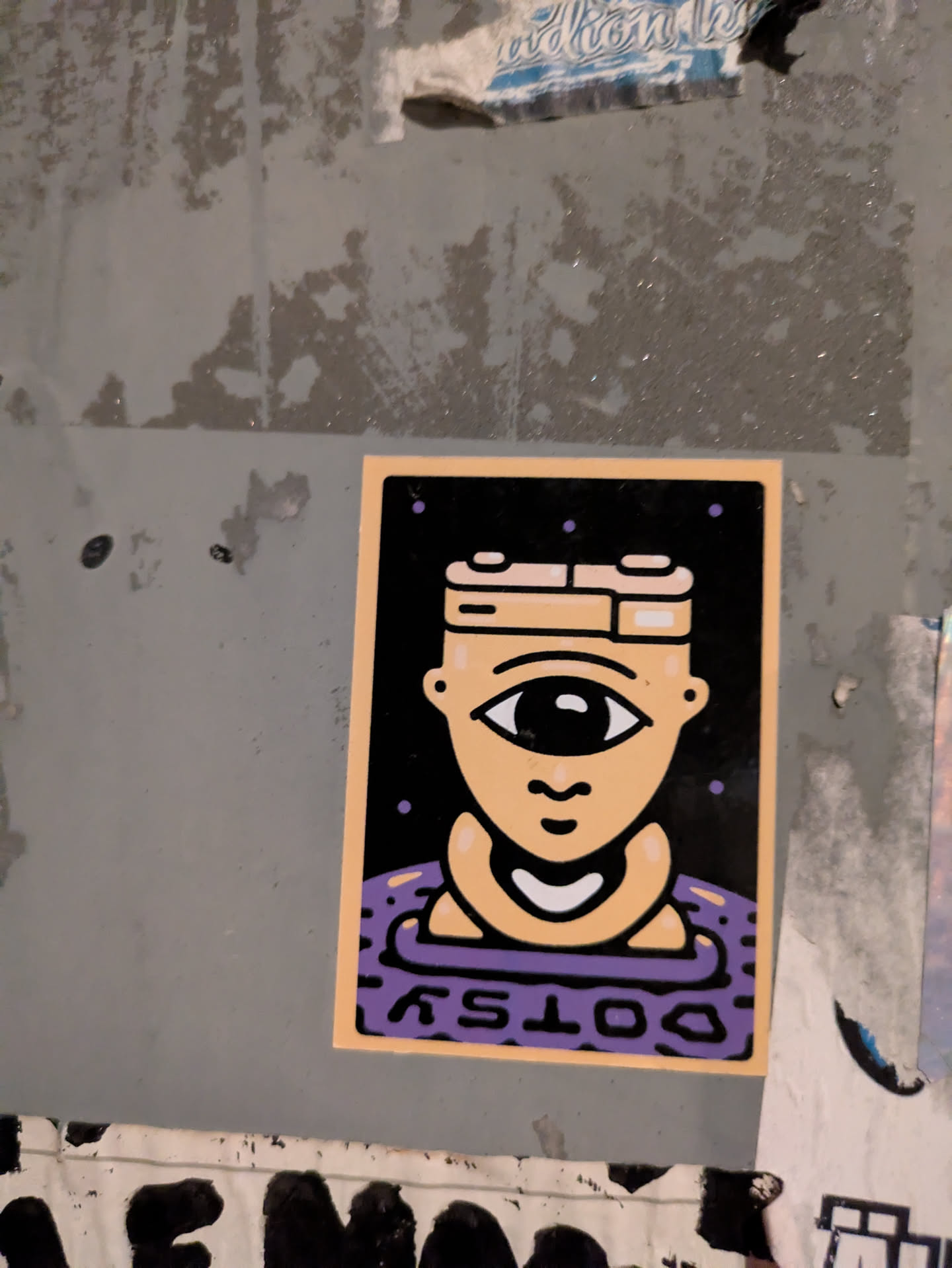 Street sticker The sticker is rectangular with a light orange-beige border, framing a central image on a black background. The main subject is a stylized, light beige head featuring a prominent single eye with a large black pupil and a small white highlight, and a simple, curved mouth below it. The head wears a distinctive, multi-layered headpiece in beige and white. The figure appears to be emerging from or submerged in a purple, wavy, or patterned surface at the bottom. Small purple dots are scattered across the black background above the head. The word "DOTSY" is written in white, inverted, across the bottom of the purple section. The overall aesthetic is clean, graphic, and illustrative.