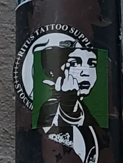 A sticker from Ritūs Tattoo Supply in Stockholm. The design depicts a person's face partially obscured by a dark object, with their middle finger raised. The background is green and the sticker has a circular border with the shop's name.