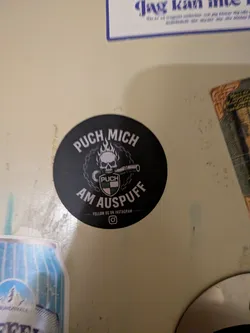 Street sticker A circular black sticker featuring a central graphic of a flaming skull biting down on a metal exhaust pipe. Below the skull is a shield emblem containing the Puch brand name and its green and white checkered logo. The central graphic is encircled by a chain border. Arched text at the top reads 'PUCH MICH' and at the bottom 'AM AUSPUFF'. Below this, the text 'FOLLOW US ON INSTAGRAM' is printed above an Instagram logo icon.