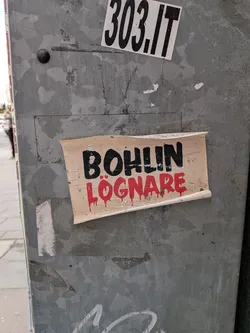 The central sticker is a rectangular piece of aged, light beige paper or vinyl, with visibly curled and peeling edges, particularly at the bottom right and top left. The background is an off-white or light tan color, showing signs of wear and dirt. The text 'BOHLIN' is prominently displayed in the upper half in thick, black, sans-serif block letters. Below it, the word 'LÖGNARE' is written in a vibrant red, stylized font that appears to be dripping, resembling blood or paint running down. The overall aesthetic is raw and impactful, suggesting a protest or strong opinion.