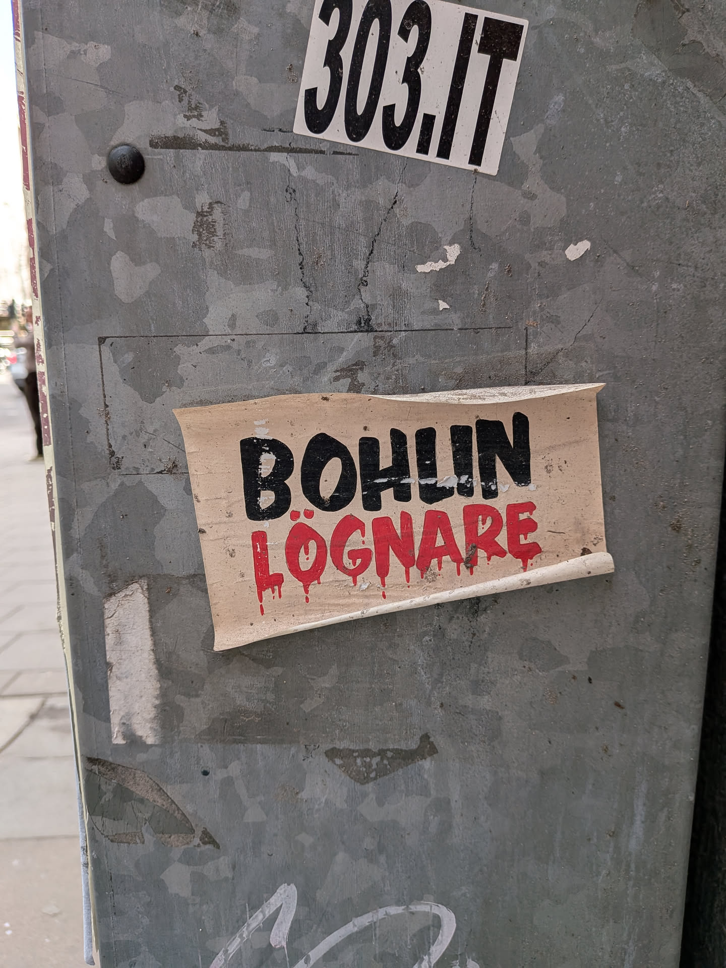 The central sticker is a rectangular piece of aged, light beige paper or vinyl, with visibly curled and peeling edges, particularly at the bottom right and top left. The background is an off-white or light tan color, showing signs of wear and dirt. The text 'BOHLIN' is prominently displayed in the upper half in thick, black, sans-serif block letters. Below it, the word 'LÖGNARE' is written in a vibrant red, stylized font that appears to be dripping, resembling blood or paint running down. The overall aesthetic is raw and impactful, suggesting a protest or strong opinion.