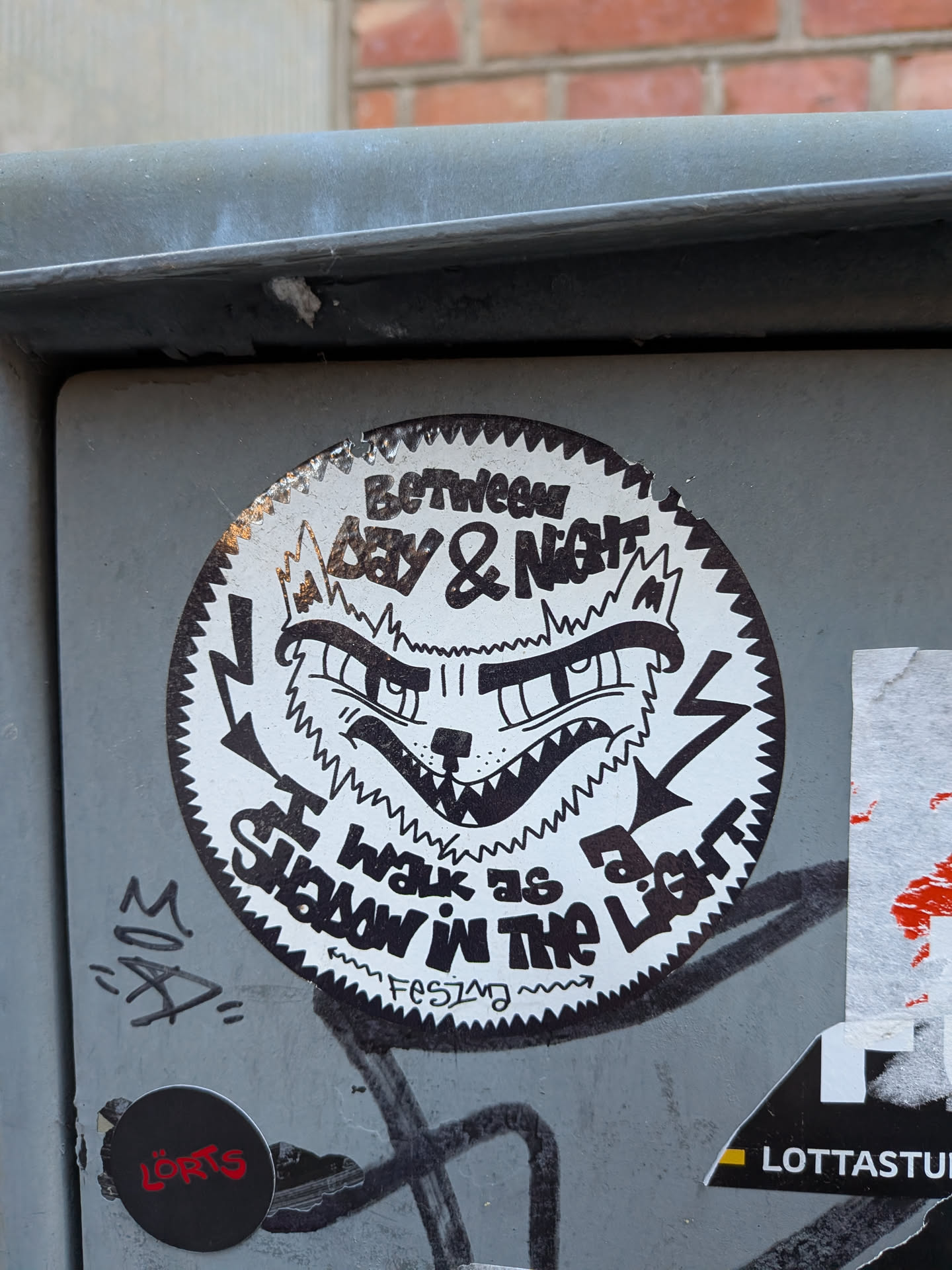 The sticker is circular, predominantly black and white, featuring a distinctive jagged, saw-tooth border. At its center is a highly stylized, aggressive-looking animal face, resembling a wolf or a fierce cat, rendered with thick, bold black lines. The face displays sharp, pointed teeth, an open mouth, an angular snout, and intense, narrowed eyes with prominent, furrowed brows. Two lightning bolt symbols are positioned on either side of the face, pointing inwards. Text is arranged in an arc around the central image: "BETWEEN DAY & NIGHT" at the top and "I SHALL WALK AS A SHADOW IN THE LIGHT" at the bottom, both in a bold, somewhat distressed, hand-drawn font. A smaller, wavy text "FESTIM" is visible below the main text at the bottom. The overall aesthetic is raw, edgy, and somewhat menacing.