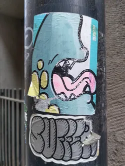 A sticker featuring a cartoon monster with a big mouth and tongue sticking out. The style is playful and colorful. Below the monster, the word "SURE" is written in stylized lettering. The sticker appears to be attached to a dark gray pole.
