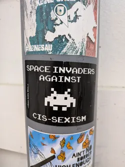 Street sticker Black rectangular sticker with white pixel art. Features a Space Invader alien between the text 'SPACE INVADERS AGAINST CIS-SEXISM'. Small vertical text on the left reads ':::Rosa Antifa Wien.::'.