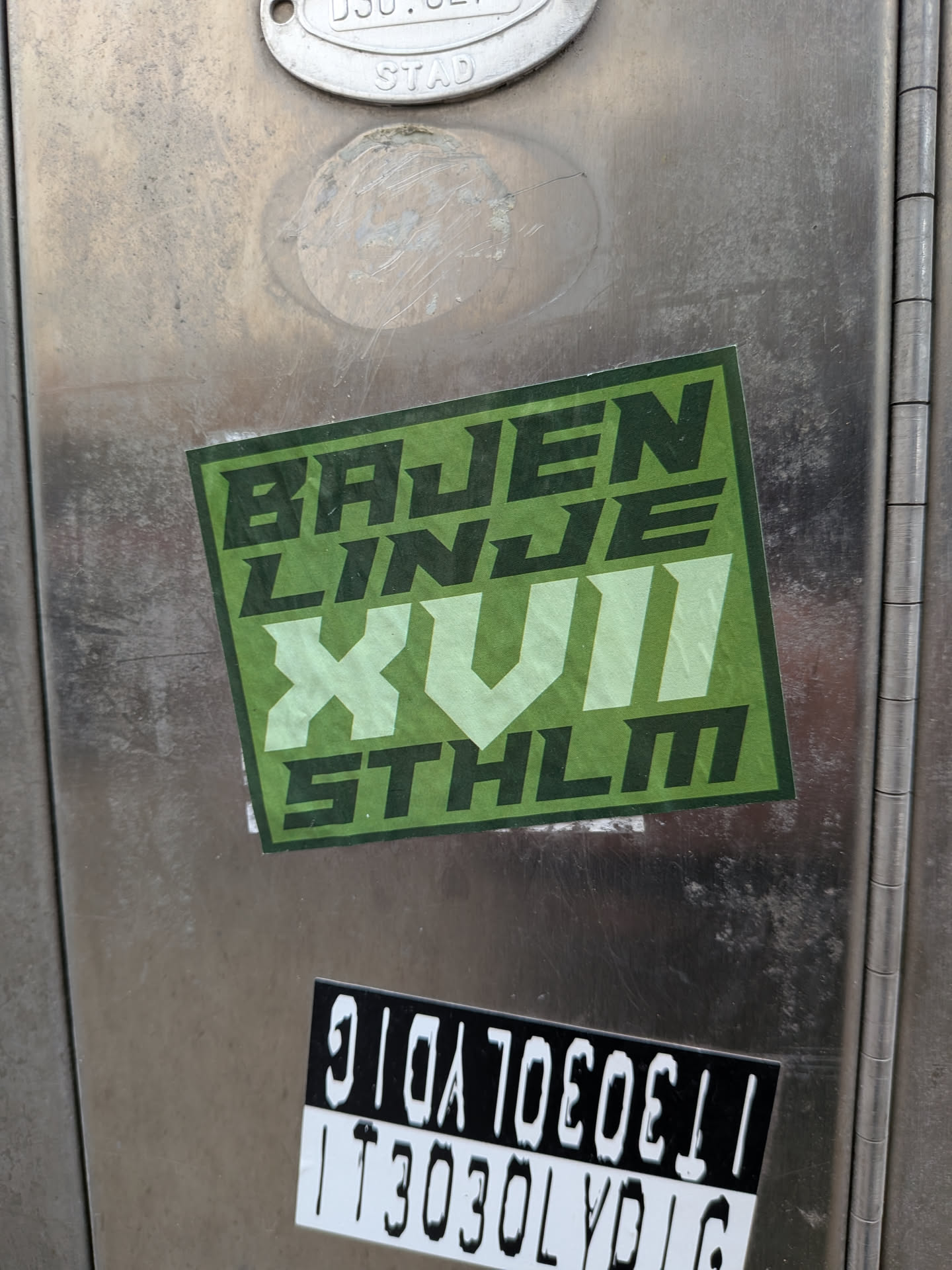 A rectangular sticker with a green color scheme, affixed to a metallic surface. The sticker features bold, blocky typography in dark and light green shades. The text is arranged in four lines: 'BAJEN' on the first, 'LINJE' on the second, a large 'XVII' (Roman numeral for 17) on the third, and 'STHLM' on the fourth.