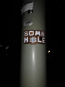Street sticker A rectangular, die-cut sticker featuring the words 'BOMB HOLE' in a prominent, bold, blocky font. The letters are white with a distinct orange outline and a black drop shadow or border. The word 'BOMB' is on the top line, and 'HOLE' is on the bottom line. The 'O' in 'HOLE' is replaced by a stylized, black and white wireframe globe or sphere. The sticker is applied to a light grey utility pole.