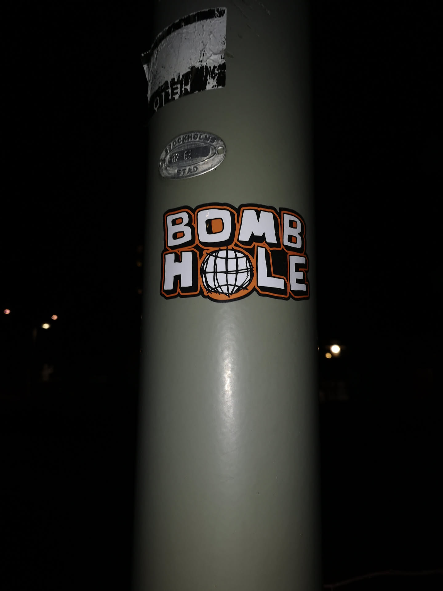 A rectangular, die-cut sticker featuring the words 'BOMB HOLE' in a prominent, bold, blocky font. The letters are white with a distinct orange outline and a black drop shadow or border. The word 'BOMB' is on the top line, and 'HOLE' is on the bottom line. The 'O' in 'HOLE' is replaced by a stylized, black and white wireframe globe or sphere. The sticker is applied to a light grey utility pole.